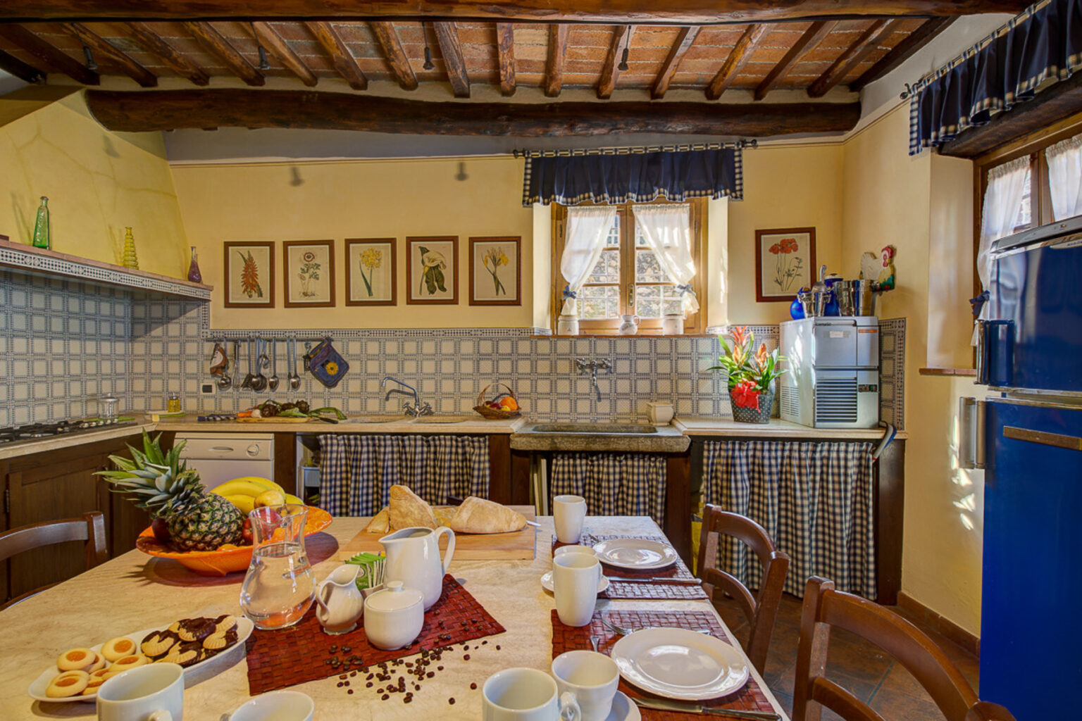 Cozy rustic kitchen with wooden beams, featuring a modern stove, a variety of fresh fruits, and a beautifully set dining table with coffee cups and pastries. Decorated with floral artwork and checkered curtains, the space offers a warm and inviting atmosphere perfect for family meals. | Only on SopranoVillas