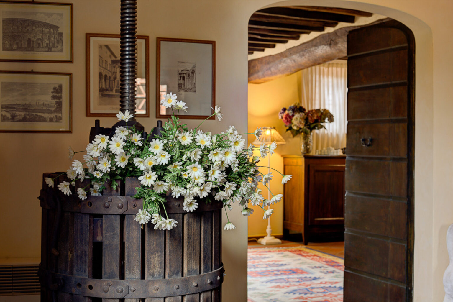 Rustic interior featuring a vintage wine press adorned with white daisies, framed artwork on the walls, and a glimpse of a warmly lit room with floral decor and a wooden cabinet in the background. | Only on SopranoVillas