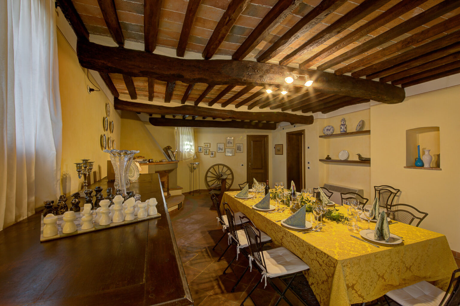 Rustic dining room with wooden beams, featuring a long table set with a yellow tablecloth and elegant tableware, surrounded by wooden chairs and decorative shelves. Natural light streams through a window, creating a warm ambiance. | Only on SopranoVillas