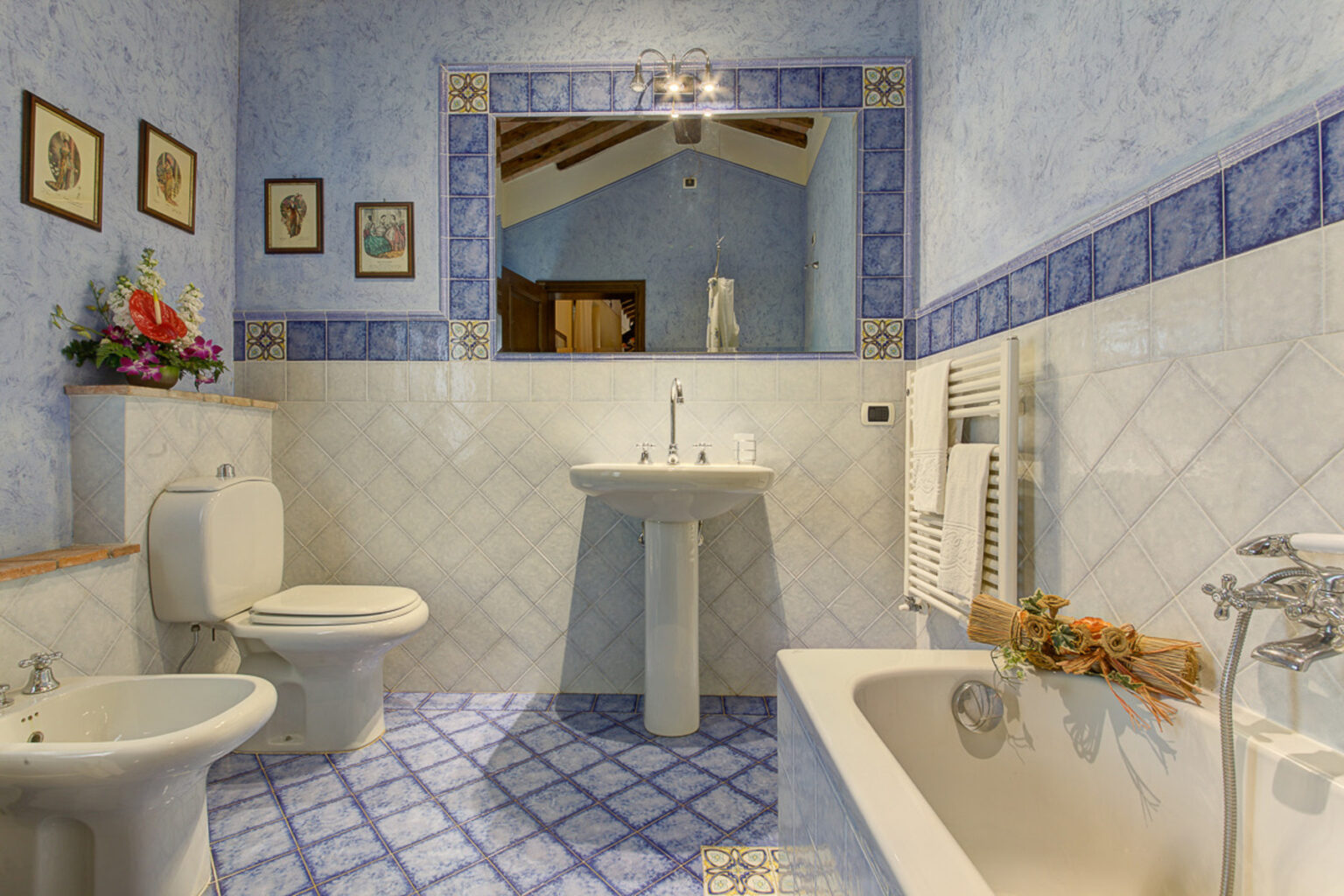 Spacious bathroom featuring blue patterned tiles, a modern sink, toilet, bidet, and a bathtub, complemented by decorative wall art and a large mirror. | Only on SopranoVillas