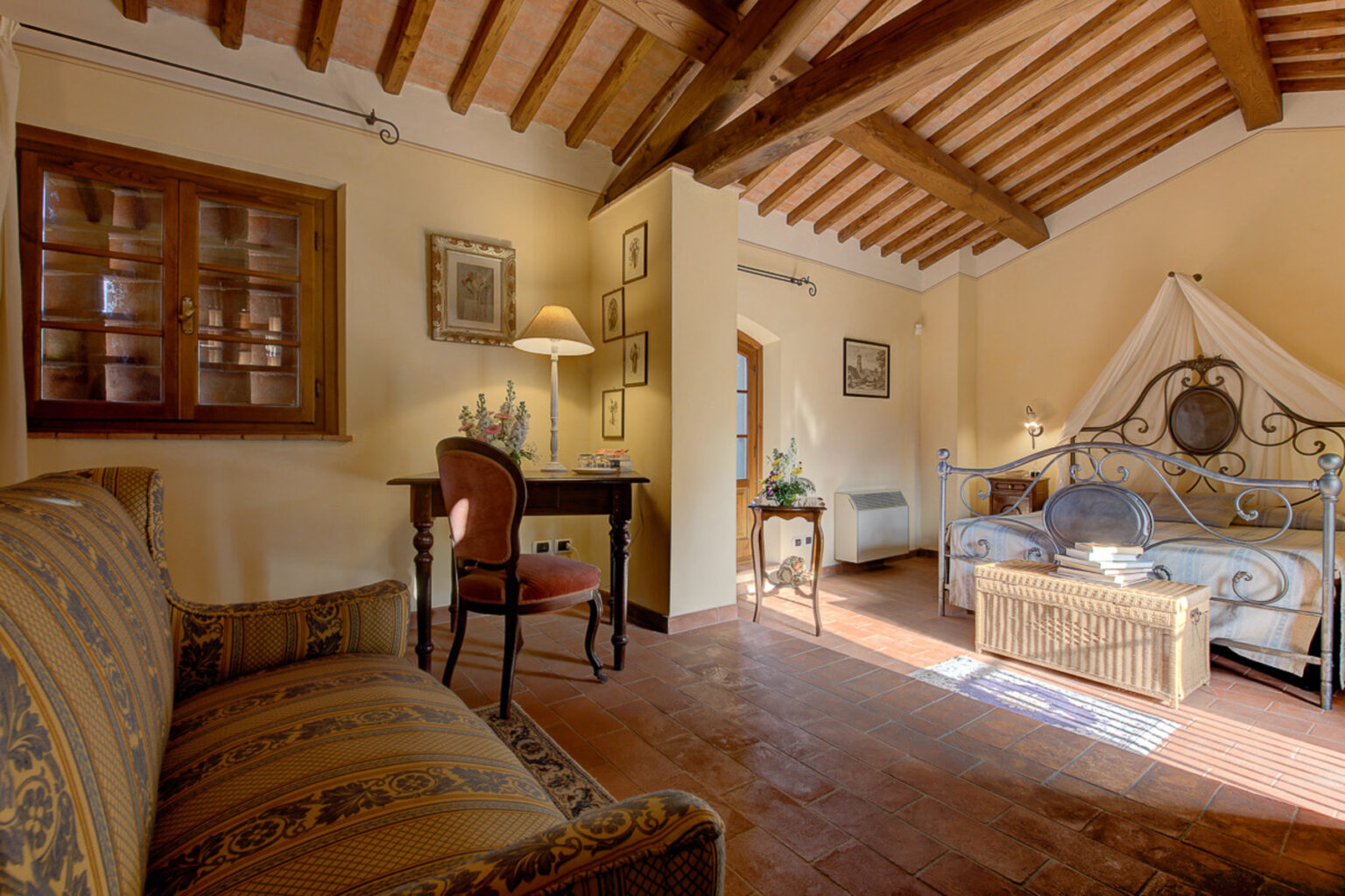 Cozy bedroom interior featuring a comfortable bed with a decorative canopy, a vintage writing desk, and elegant furnishings. Natural light illuminates the warm, rustic decor, highlighting wooden beams and charming artwork on the walls. Ideal for a relaxing getaway. | Only on SopranoVillas