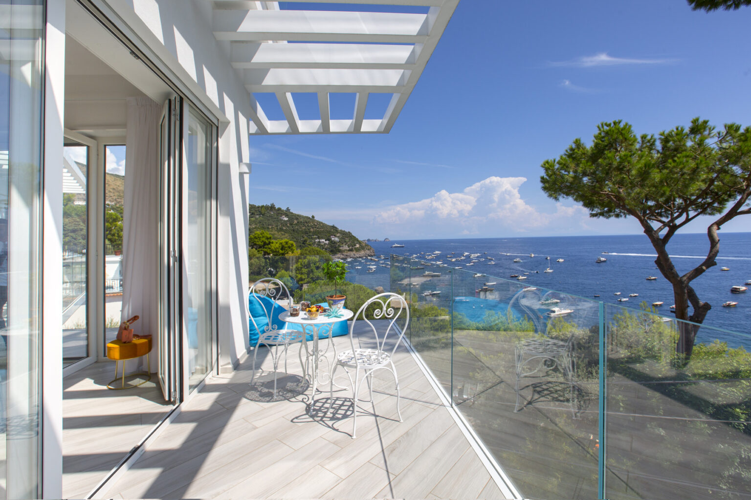 Modern balcony with glass railing overlooking a serene coastal view, featuring a table set for breakfast, surrounded by lush greenery and blue waters under a clear sky. | Only on SopranoVillas