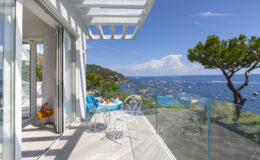 Modern balcony with glass railing overlooking a serene coastal view, featuring a table set for breakfast, surrounded by lush greenery and blue waters under a clear sky.