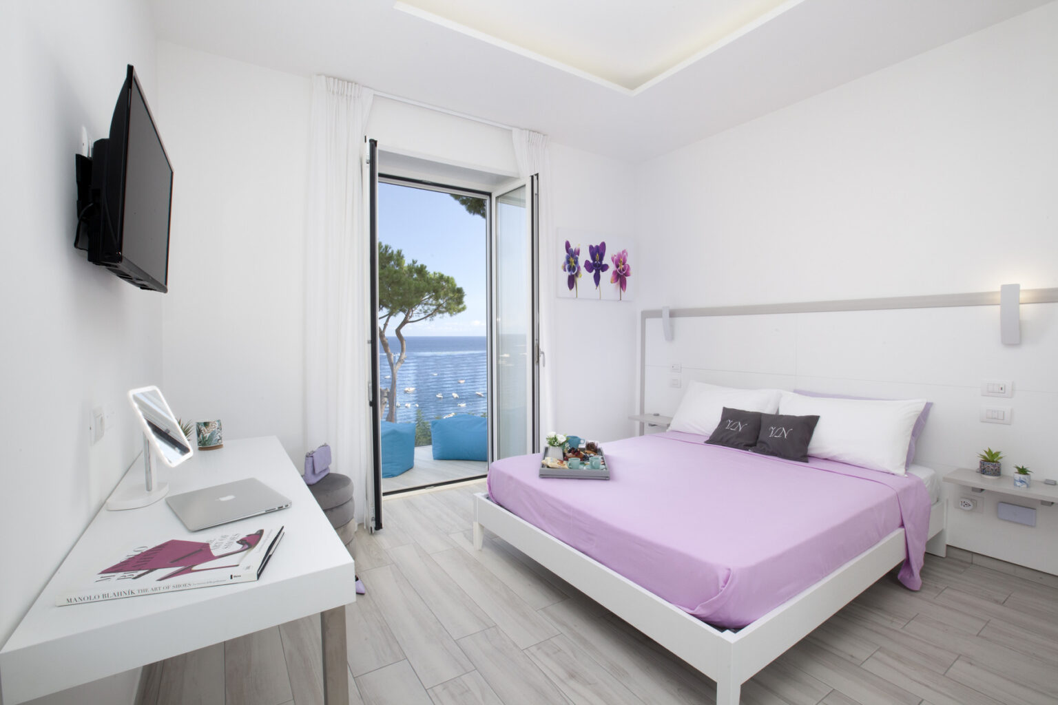 Modern hotel room with a purple bedspread, a wall-mounted TV, and a stylish desk. The room features large windows offering a view of the sea and outdoor seating. Bright, minimalist decor with floral artwork enhances the serene atmosphere. | Only on SopranoVillas