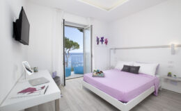Modern hotel room with a purple bedspread, a wall-mounted TV, and a stylish desk. The room features large windows offering a view of the sea and outdoor seating. Bright, minimalist decor with floral artwork enhances the serene atmosphere.