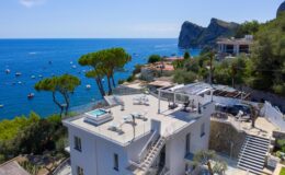 Luxury coastal villa with a rooftop terrace, overlooking the blue sea and boats, surrounded by lush greenery and rocky cliffs. Ideal for relaxation and stunning views.