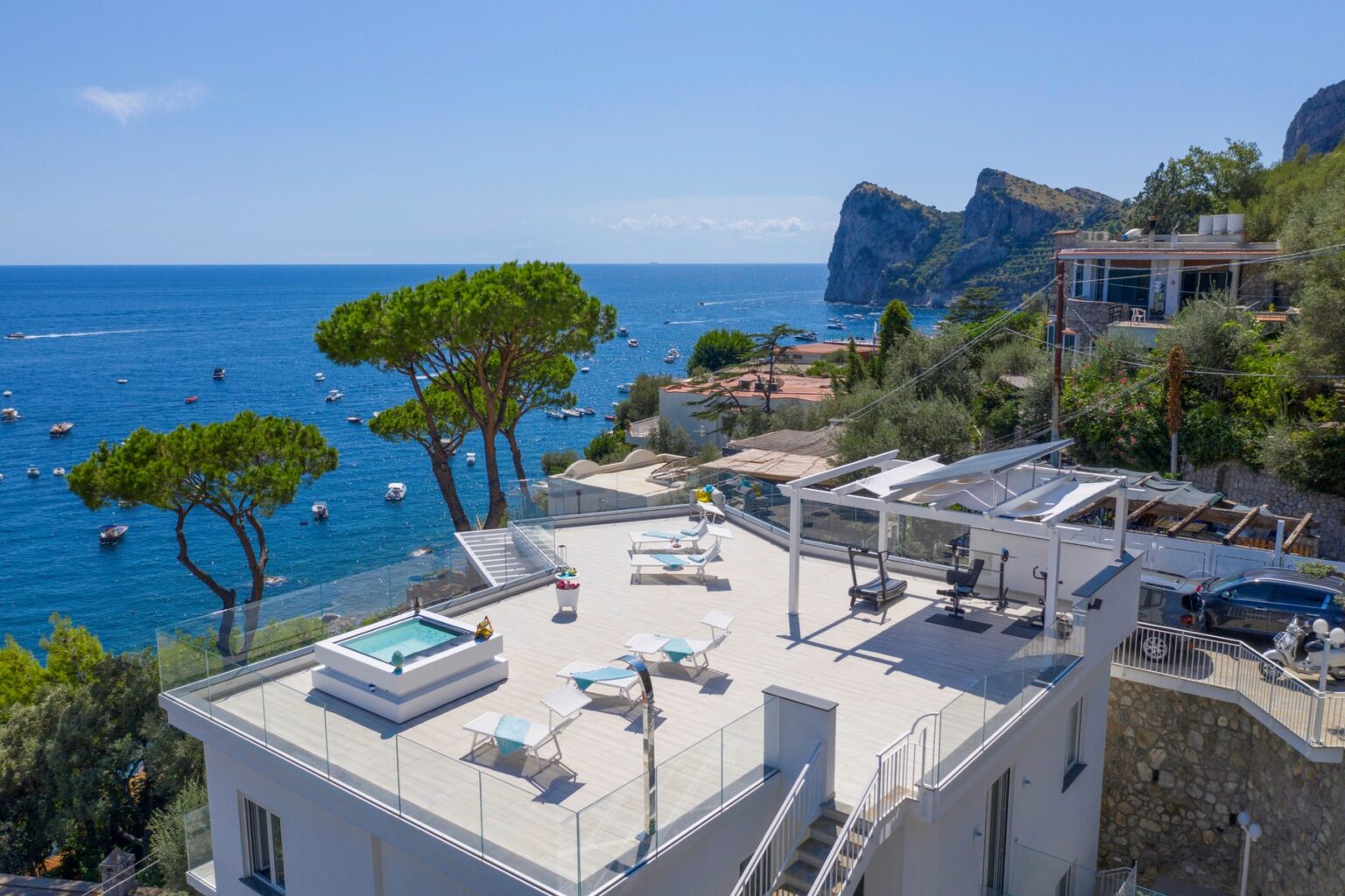 Modern villa with a rooftop terrace featuring a hot tub and lounge chairs, overlooking the blue sea and boats. Lush pine trees and coastal scenery enhance the luxurious ambiance. Ideal for relaxation and stunning views. | Only on SopranoVillas