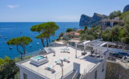 Modern villa with a rooftop terrace featuring a hot tub and lounge chairs, overlooking the blue sea and boats. Lush pine trees and coastal scenery enhance the luxurious ambiance. Ideal for relaxation and stunning views.