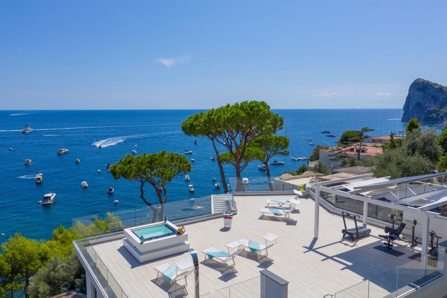 Luxury villa terrace overlooking the Mediterranean Sea, featuring a hot tub, sun loungers, and a panoramic view of boats and coastal scenery. | Only on SopranoVillas