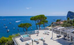 Luxury villa terrace overlooking the Mediterranean Sea, featuring a hot tub, sun loungers, and a panoramic view of boats and coastal scenery.
