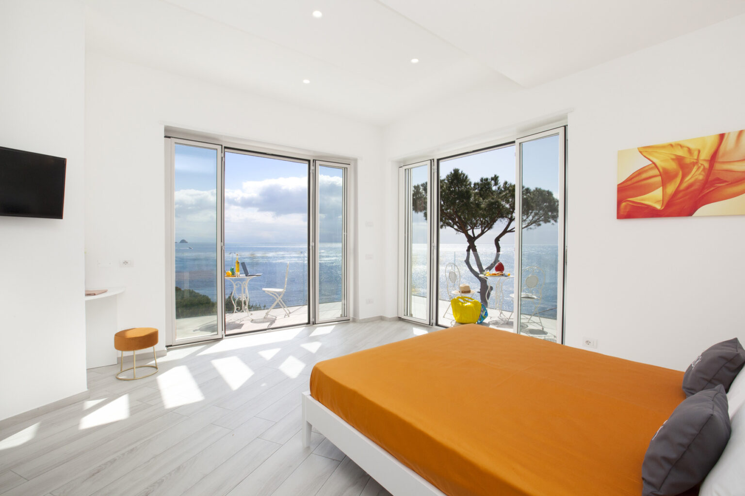 Modern bedroom with an orange bedspread, large windows offering a scenic view of the ocean and landscape, minimalist decor, and a bright atmosphere. Ideal for relaxation and comfort in a coastal setting. | Only on SopranoVillas
