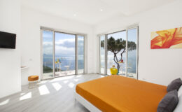 Modern bedroom with an orange bedspread, large windows offering a scenic view of the ocean and landscape, minimalist decor, and a bright atmosphere. Ideal for relaxation and comfort in a coastal setting.