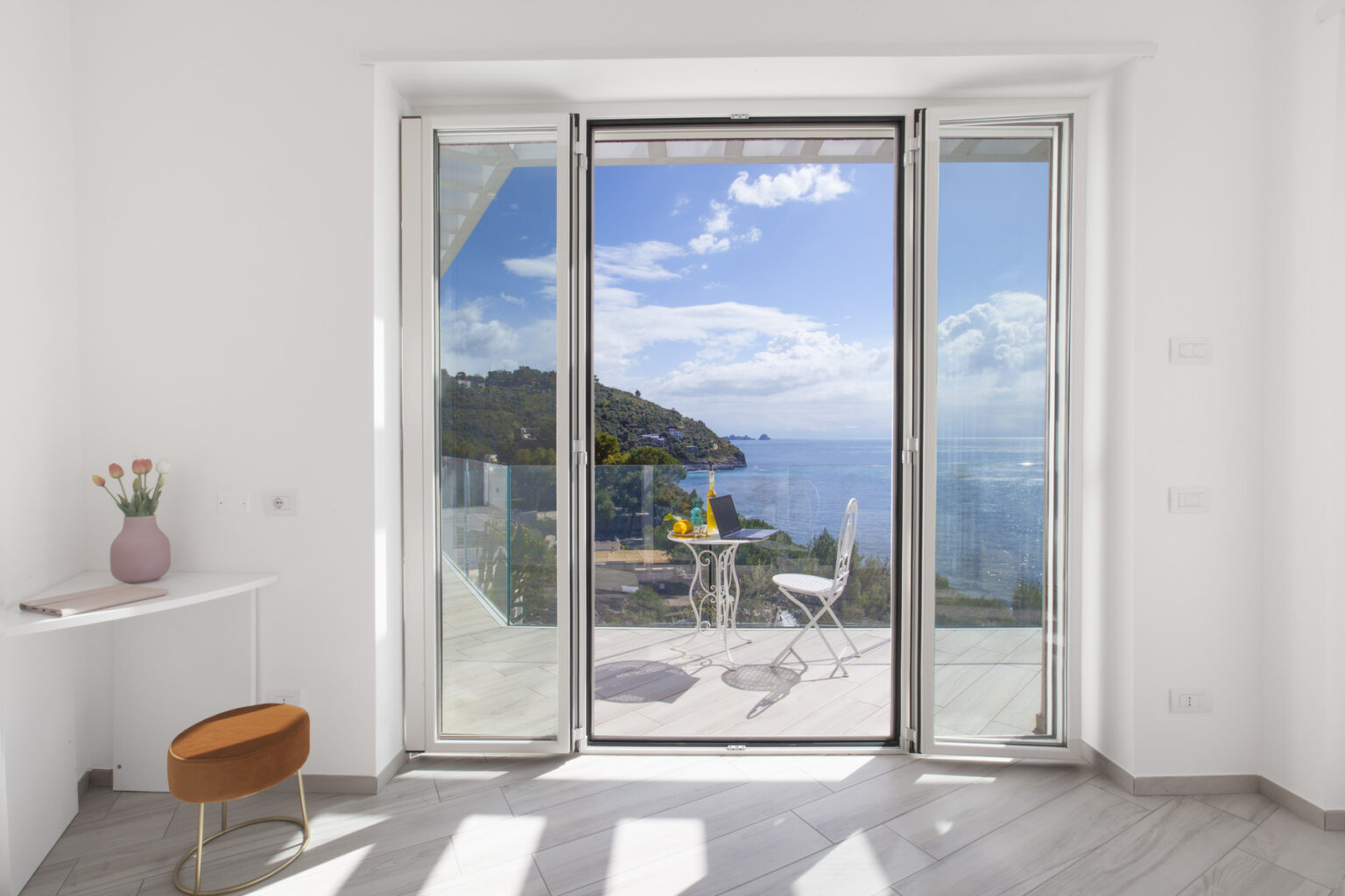 Modern interior with large glass doors opening to a scenic ocean view, featuring a small table and chairs on a balcony, surrounded by lush greenery and blue skies. | Only on SopranoVillas