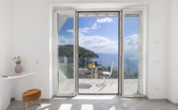 Modern interior with large glass doors opening to a scenic ocean view, featuring a small table and chairs on a balcony, surrounded by lush greenery and blue skies.