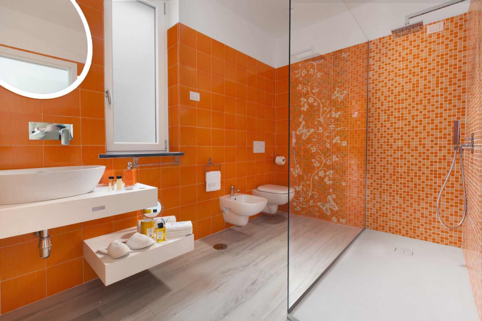 Modern bathroom featuring vibrant orange wall tiles, a glass shower enclosure, a sleek white sink with toiletries, and contemporary fixtures. | Only on SopranoVillas