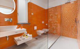 Modern bathroom featuring vibrant orange wall tiles, a glass shower enclosure, a sleek white sink with toiletries, and contemporary fixtures.