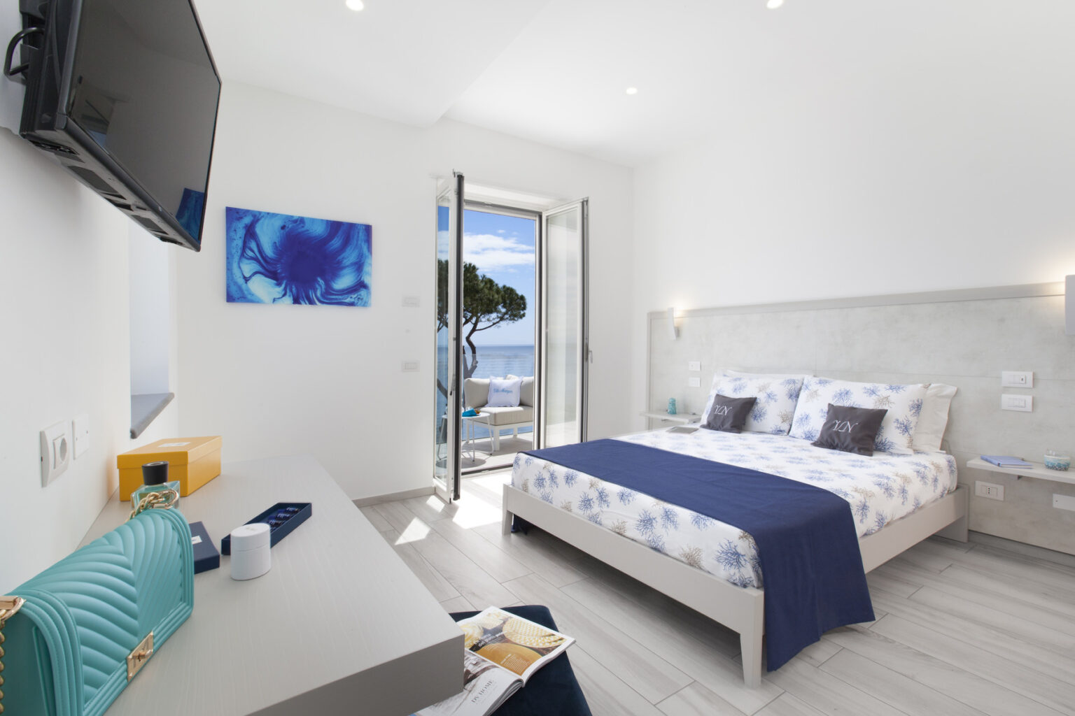 Modern hotel room with a double bed, featuring white and blue decor, a flat-screen TV, and a desk. The room has large windows that open to a scenic view, allowing natural light to fill the space. Ideal for travelers seeking comfort and beautiful surroundings. | Only on SopranoVillas