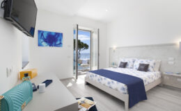Modern hotel room with a double bed, featuring white and blue decor, a flat-screen TV, and a desk. The room has large windows that open to a scenic view, allowing natural light to fill the space. Ideal for travelers seeking comfort and beautiful surroundings.
