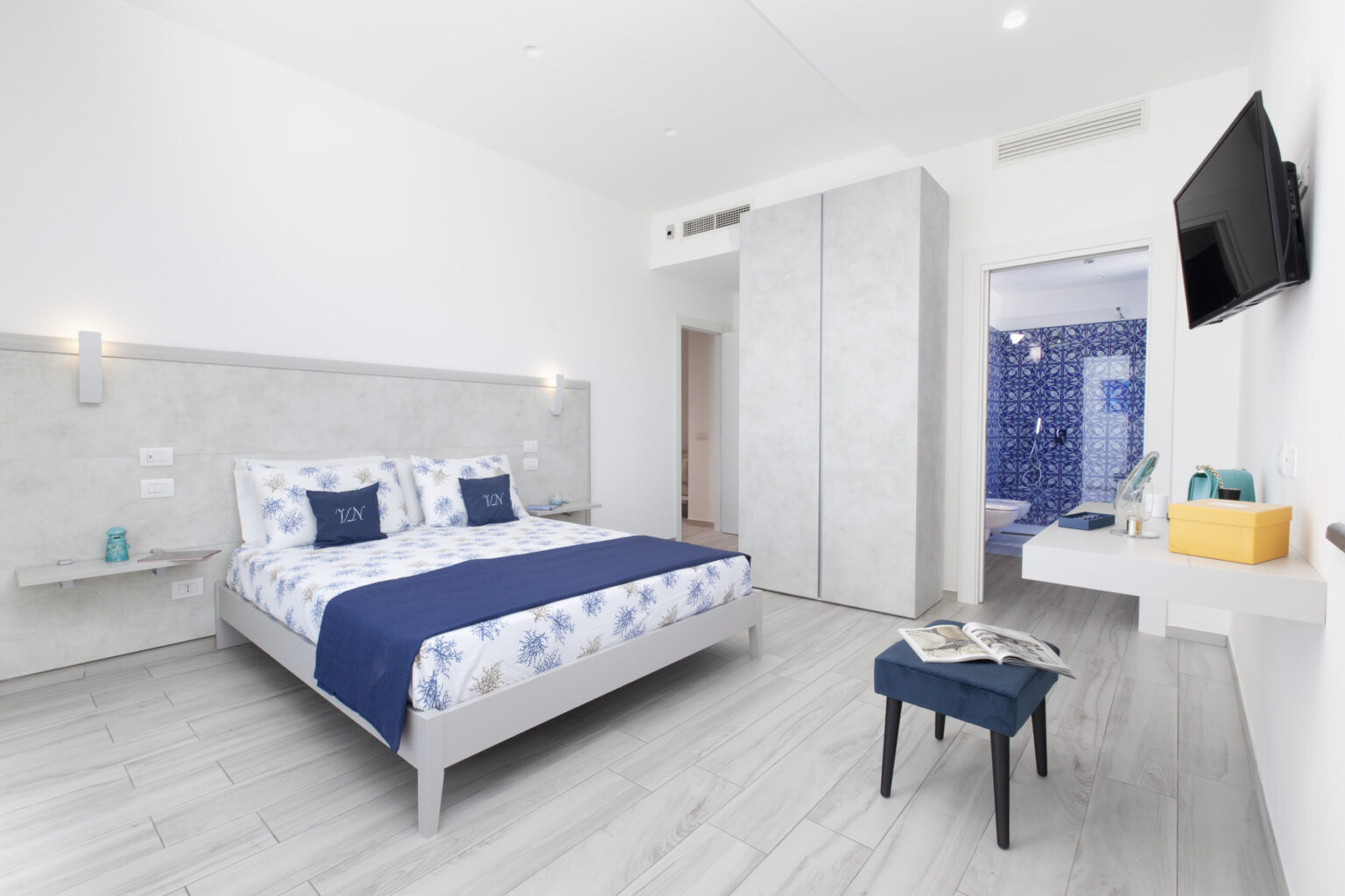 Modern hotel room featuring a king-size bed with blue and white bedding, minimalist decor, wooden flooring, a wall-mounted TV, and an en-suite bathroom. Ideal for travelers seeking comfort and style. | Only on SopranoVillas