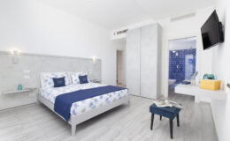 Modern hotel room featuring a king-size bed with blue and white bedding, minimalist decor, wooden flooring, a wall-mounted TV, and an en-suite bathroom. Ideal for travelers seeking comfort and style.