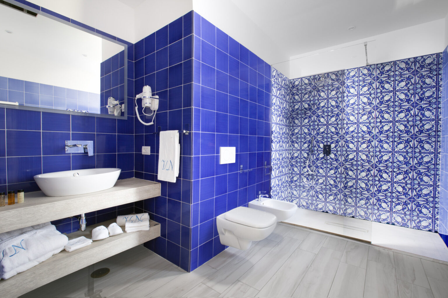 Modern bathroom featuring vibrant blue tiled walls, a stylish vessel sink on a wooden vanity, and a contemporary shower area with intricate patterned tiles. Soft towels and essential toiletries are neatly arranged, enhancing the aesthetic appeal. | Only on SopranoVillas