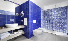 Modern bathroom featuring vibrant blue tiled walls, a stylish vessel sink on a wooden vanity, and a contemporary shower area with intricate patterned tiles. Soft towels and essential toiletries are neatly arranged, enhancing the aesthetic appeal.
