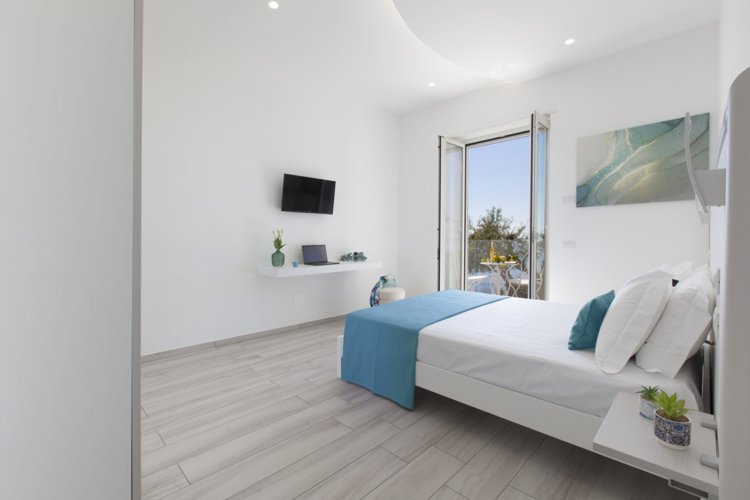 Modern bedroom featuring a white bed with blue accents, a wall-mounted TV, and a workspace. The room has large windows with a view, allowing natural light to fill the space. Decor includes a vase with flowers and small plants on bedside tables, creating a fresh and inviting atmosphere. | Only on SopranoVillas