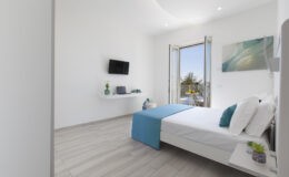 Modern bedroom featuring a white bed with blue accents, a wall-mounted TV, and a workspace. The room has large windows with a view, allowing natural light to fill the space. Decor includes a vase with flowers and small plants on bedside tables, creating a fresh and inviting atmosphere.