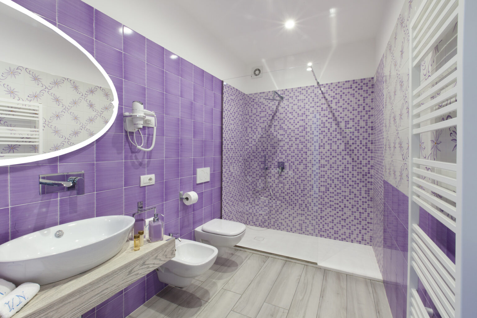 Modern bathroom design featuring purple tiled walls, a spacious walk-in shower, a sleek oval mirror, and contemporary fixtures. The room includes a stylish sink and bidet, complemented by warm wood accents and efficient lighting. | Only on SopranoVillas