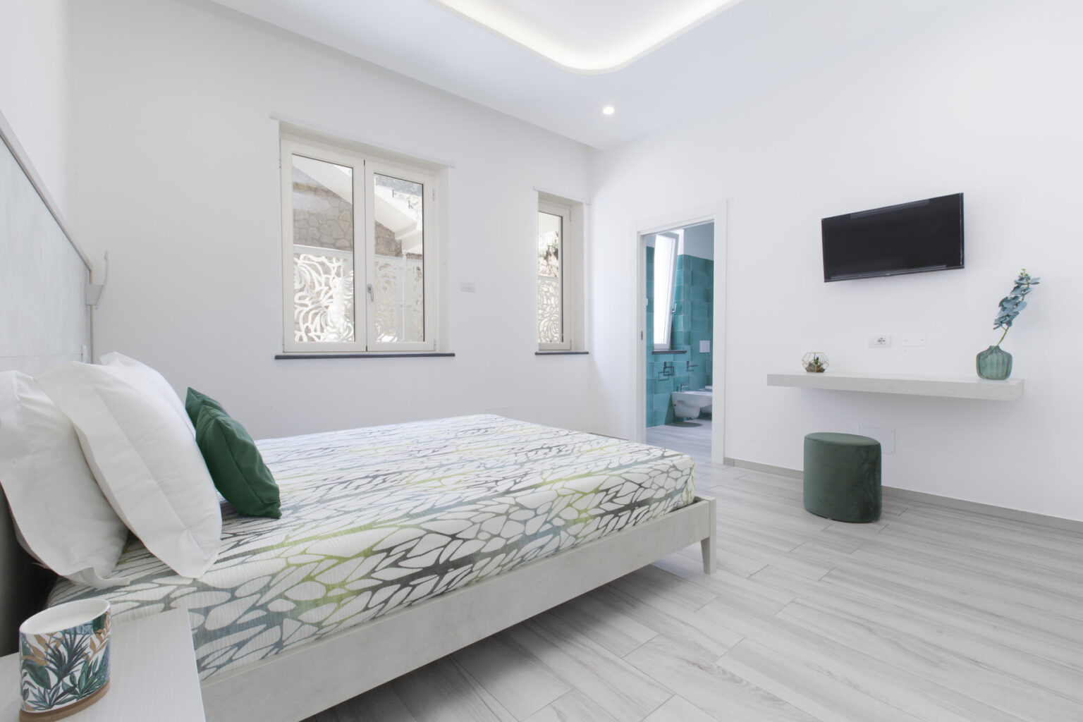 Modern bedroom featuring a queen-sized bed with a floral-patterned comforter, minimalist decor, a wall-mounted TV, and a small side table. Natural light streams through large windows, enhancing the bright and airy atmosphere. | Only on SopranoVillas