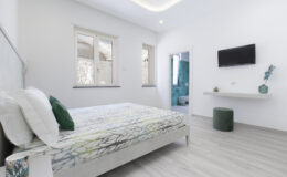 Modern bedroom featuring a queen-sized bed with a floral-patterned comforter, minimalist decor, a wall-mounted TV, and a small side table. Natural light streams through large windows, enhancing the bright and airy atmosphere.