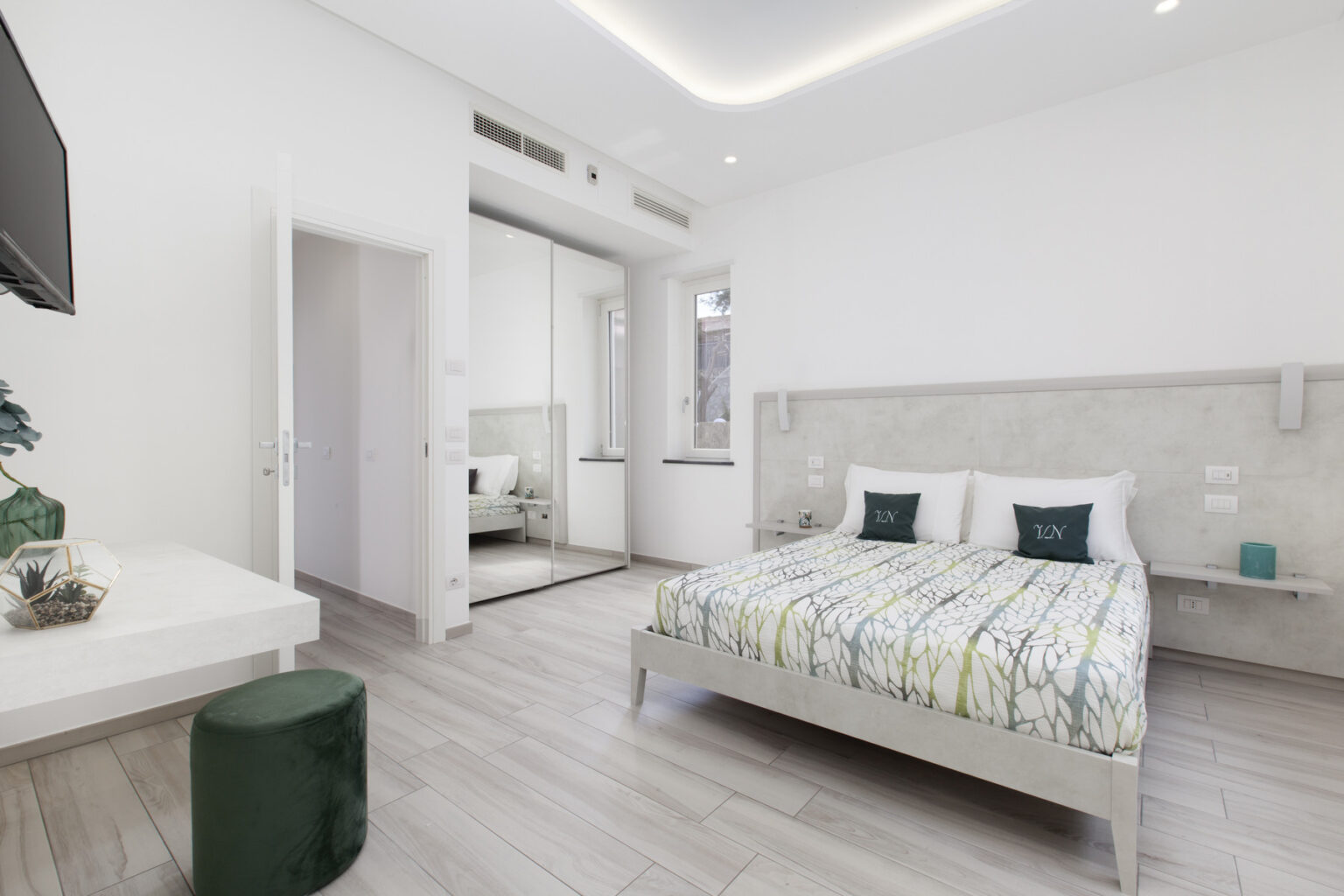 Modern bedroom featuring a comfortable bed with a patterned duvet, minimalist decor, a flat-screen TV, and natural light from large windows. The room includes a small green stool and a sleek white dresser, creating a cozy and inviting atmosphere. | Only on SopranoVillas