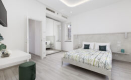 Modern bedroom featuring a comfortable bed with a patterned duvet, minimalist decor, a flat-screen TV, and natural light from large windows. The room includes a small green stool and a sleek white dresser, creating a cozy and inviting atmosphere.