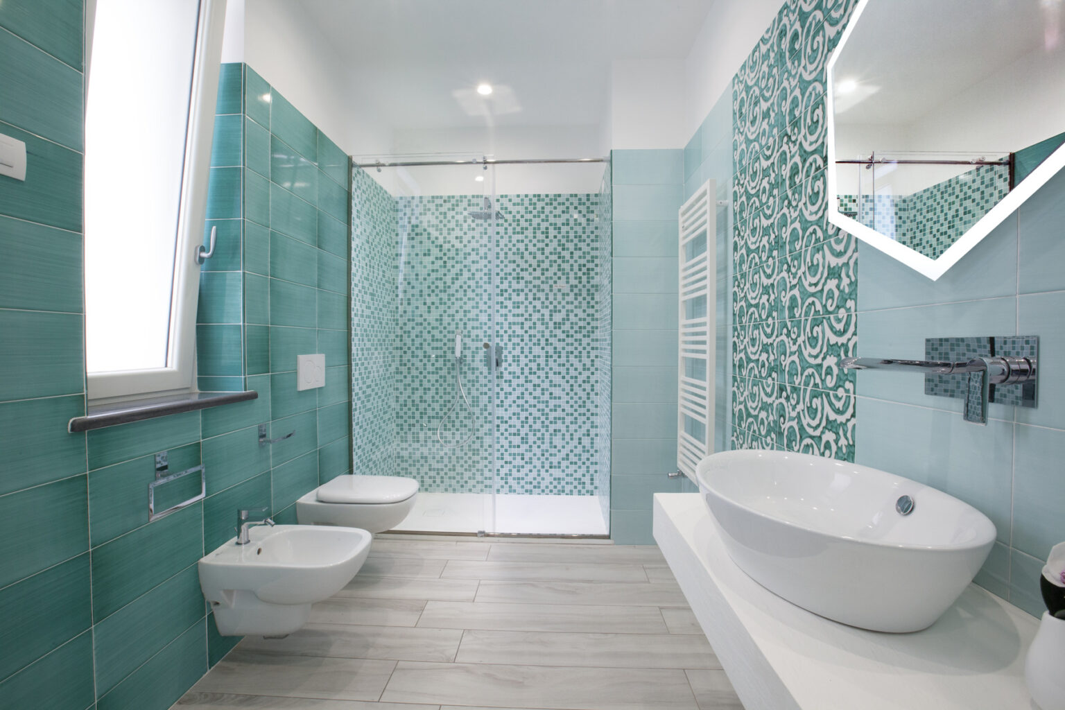 Modern bathroom design featuring turquoise patterned tiles, a freestanding bathtub, a bidet, and a walk-in shower with glass doors. The space is bright and stylish, showcasing contemporary fixtures and a geometric mirror. | Only on SopranoVillas