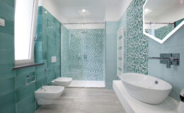 Modern bathroom design featuring turquoise patterned tiles, a freestanding bathtub, a bidet, and a walk-in shower with glass doors. The space is bright and stylish, showcasing contemporary fixtures and a geometric mirror.