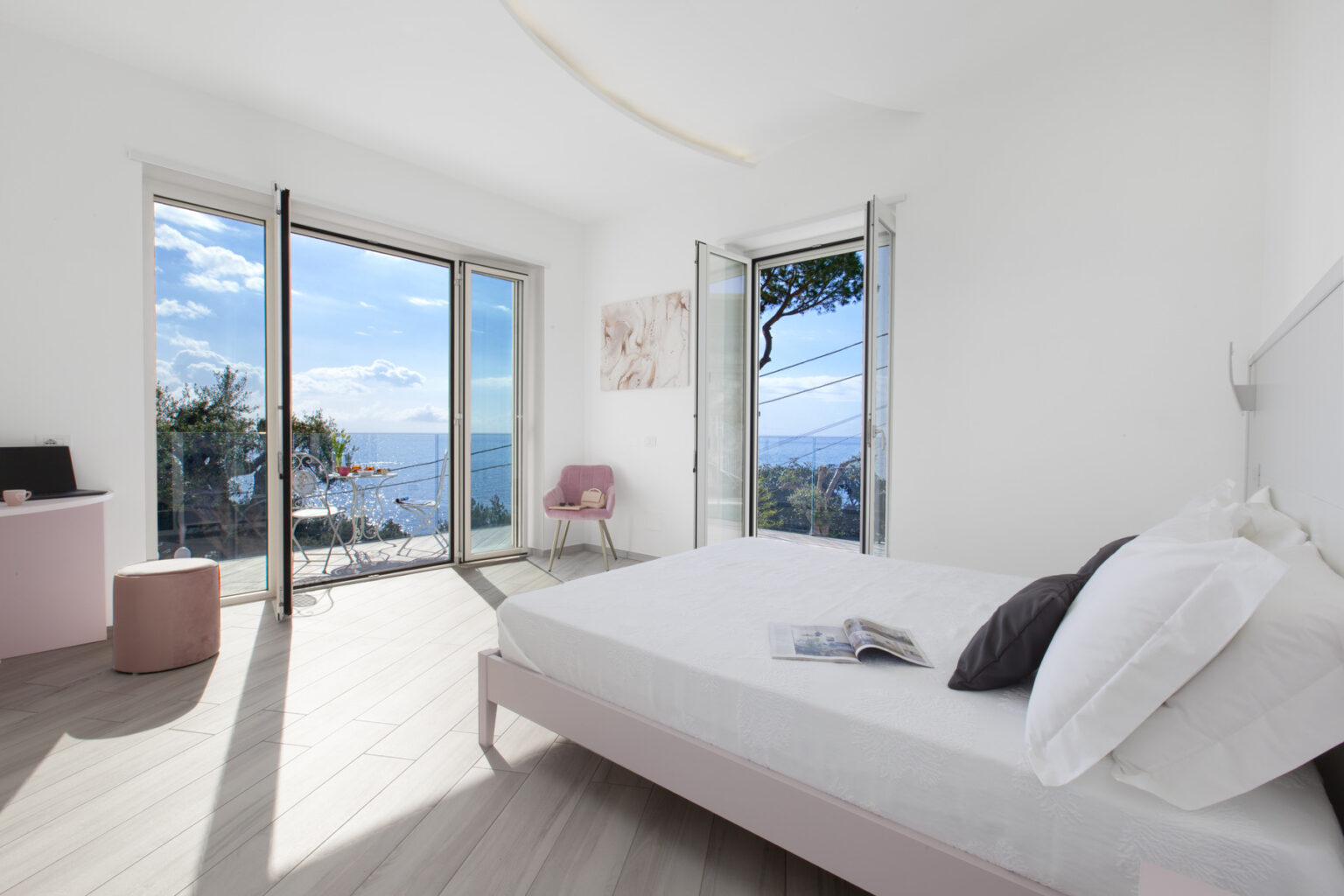 Bright and airy modern bedroom with large windows showcasing a scenic view of the ocean and landscape. The room features minimalist decor, a comfortable bed with white linens, a pink accent chair, and wooden flooring, creating a serene atmosphere. | Only on SopranoVillas
