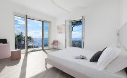 Bright and airy modern bedroom with large windows showcasing a scenic view of the ocean and landscape. The room features minimalist decor, a comfortable bed with white linens, a pink accent chair, and wooden flooring, creating a serene atmosphere.