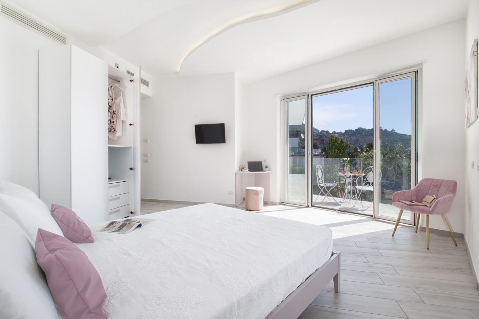 Modern bedroom with a large bed, soft pink accents, a flat-screen TV, and a sliding glass door leading to a balcony with mountain views. The room features bright white walls and light wood flooring, creating a spacious and airy atmosphere. | Only on SopranoVillas