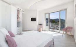 Modern bedroom with a large bed, soft pink accents, a flat-screen TV, and a sliding glass door leading to a balcony with mountain views. The room features bright white walls and light wood flooring, creating a spacious and airy atmosphere.