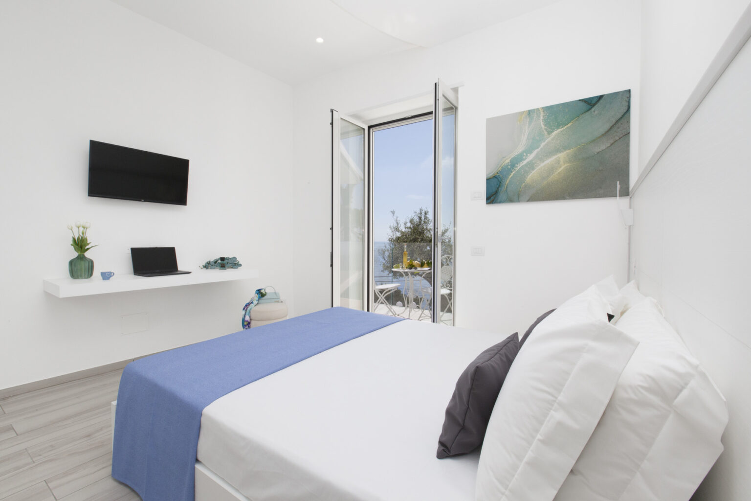 Bright and modern bedroom featuring a queen-sized bed with white linens and a blue throw, a flat-screen TV on the wall, a desk, and large windows opening to a scenic view. The room is decorated with minimalist furnishings and soft natural light. | Only on SopranoVillas
