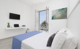 Bright and modern bedroom featuring a queen-sized bed with white linens and a blue throw, a flat-screen TV on the wall, a desk, and large windows opening to a scenic view. The room is decorated with minimalist furnishings and soft natural light.