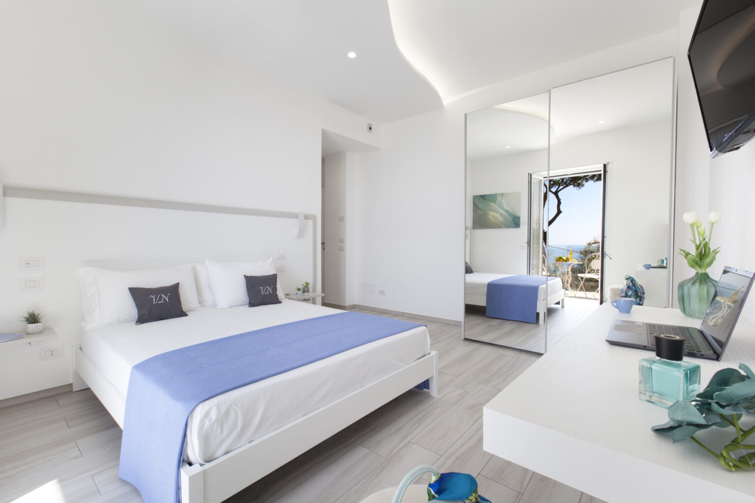 Modern hotel bedroom featuring a queen-sized bed with a blue throw, minimalist decor, a large mirror, and a view of the outdoors. The room is bright and airy, with neutral tones and contemporary furnishings. | Only on SopranoVillas