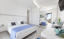 Modern hotel bedroom featuring a queen-sized bed with a blue throw, minimalist decor, a large mirror, and a view of the outdoors. The room is bright and airy, with neutral tones and contemporary furnishings.