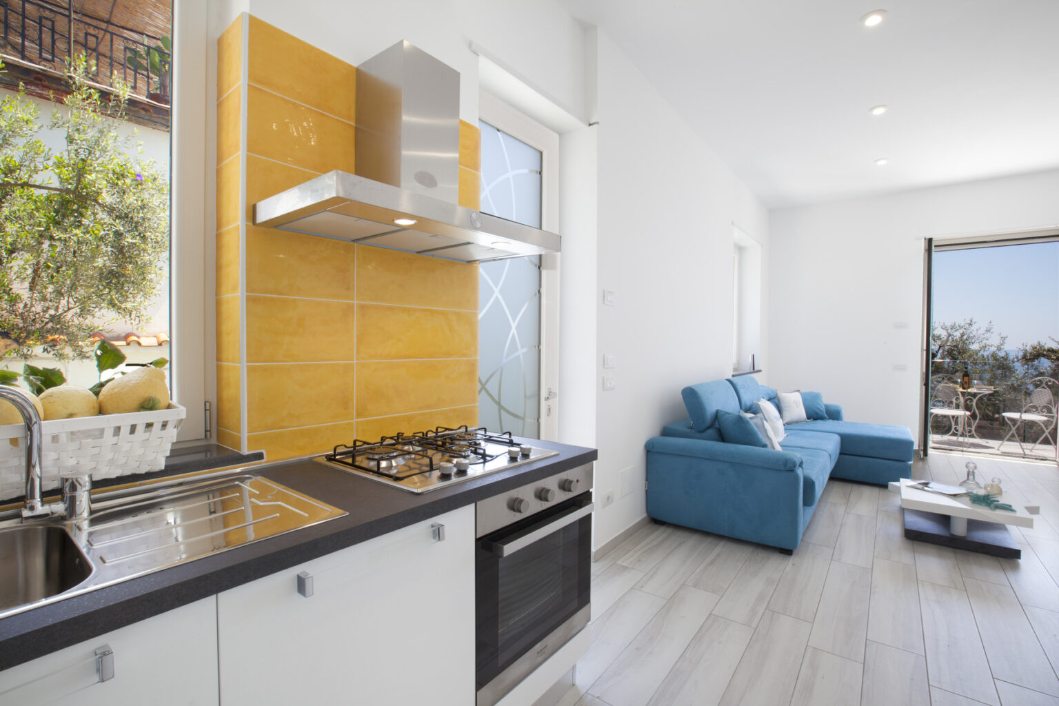 Modern kitchen with stainless steel appliances, yellow backsplash, and a cozy living area featuring a blue sofa and large windows overlooking a scenic view. | Only on SopranoVillas
