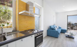 Modern kitchen with stainless steel appliances, yellow backsplash, and a cozy living area featuring a blue sofa and large windows overlooking a scenic view.