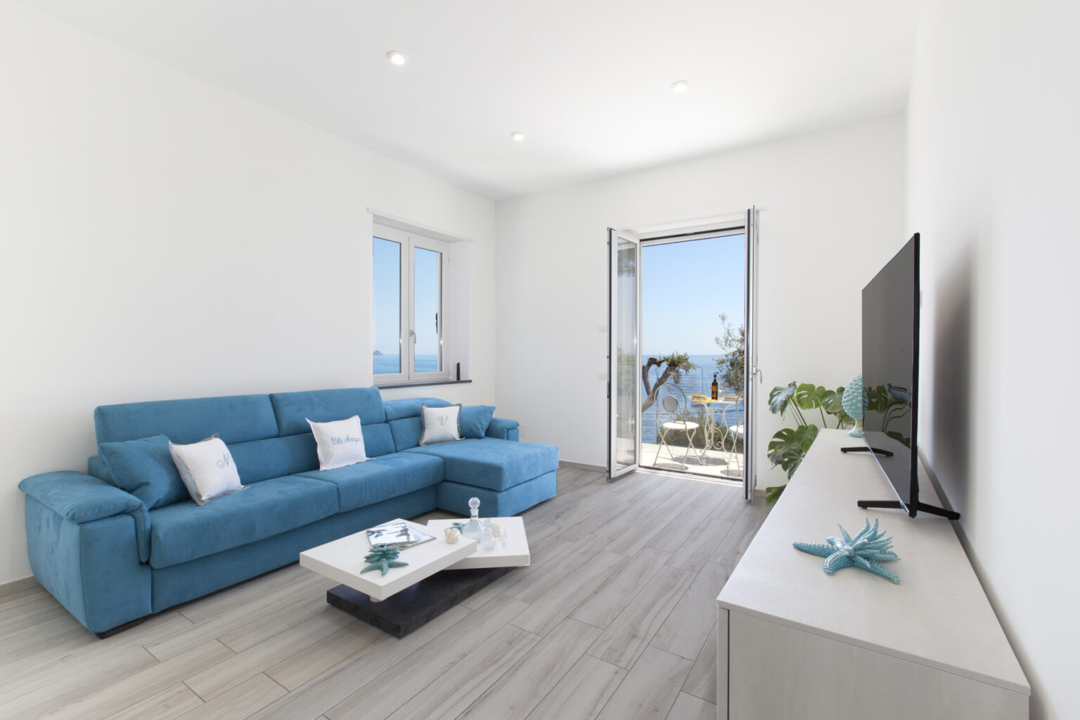 Bright and modern living room featuring a blue sofa, a coffee table, and a TV, with large windows showcasing a scenic view of the outdoors. The space has light-colored walls and wooden flooring, creating a fresh and airy atmosphere. | Only on SopranoVillas