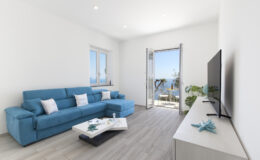 Bright and modern living room featuring a blue sofa, a coffee table, and a TV, with large windows showcasing a scenic view of the outdoors. The space has light-colored walls and wooden flooring, creating a fresh and airy atmosphere.