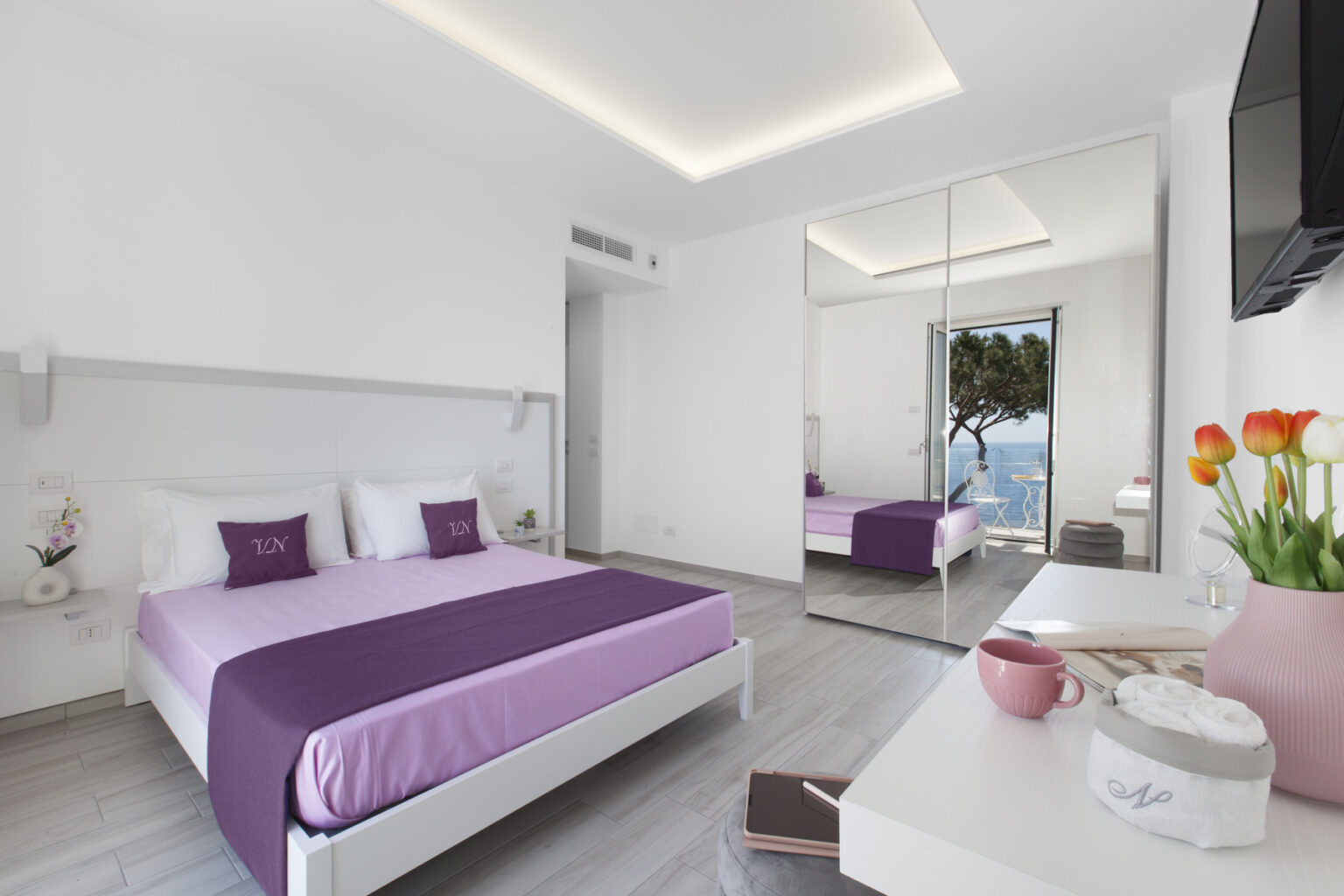 Modern hotel room featuring a purple bedspread, minimalist decor, and large windows with a scenic view. The room includes a mirrored wardrobe, a flat-screen TV, and a stylish bedside table with flowers. | Only on SopranoVillas