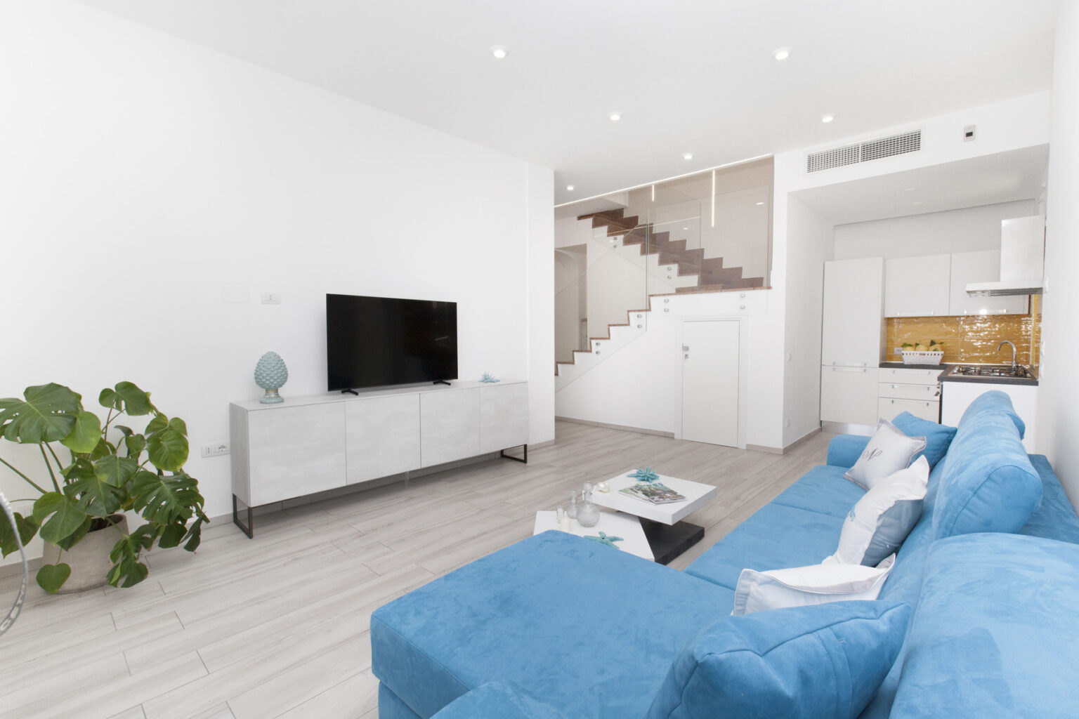 Modern living room featuring a blue sectional sofa, a sleek white entertainment unit with a flat-screen TV, and a stylish coffee table. The space includes a decorative plant and a staircase leading to an upper level, with light wood flooring and bright, airy decor. | Only on SopranoVillas