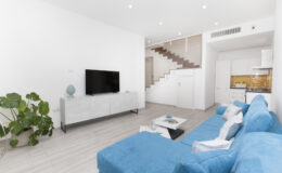 Modern living room featuring a blue sectional sofa, a sleek white entertainment unit with a flat-screen TV, and a stylish coffee table. The space includes a decorative plant and a staircase leading to an upper level, with light wood flooring and bright, airy decor.